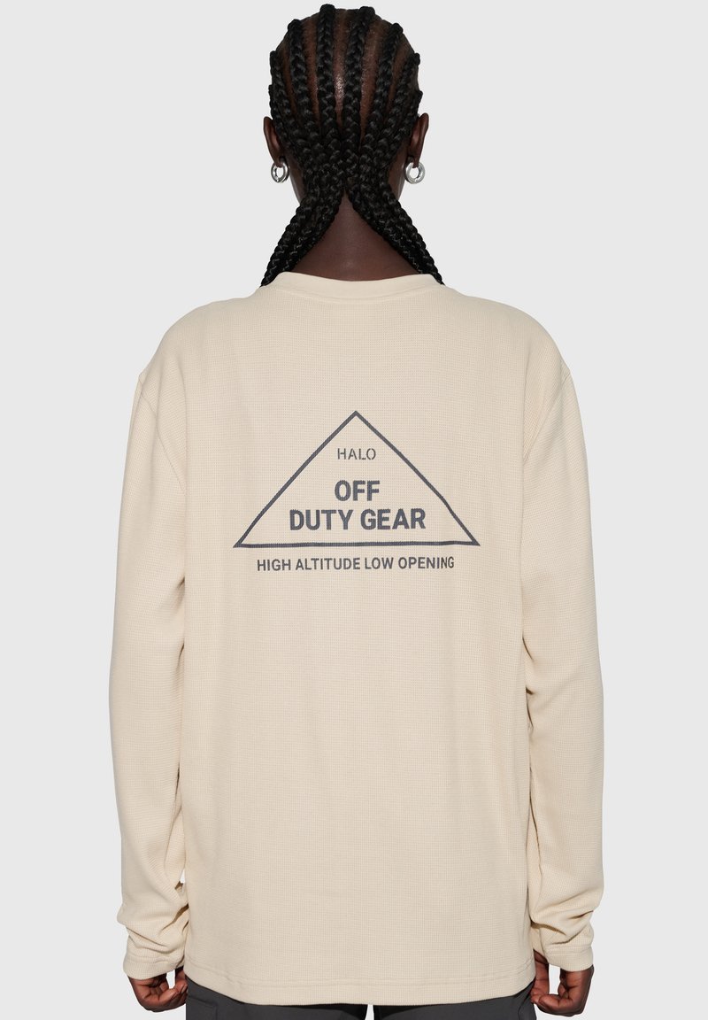 Beige långärmad topp i strukturerat tyg. Baksidan pryds av en triangelgrafik med texten: "HALO OFF DUTY GEAR HIGH ALTITUDE LOW OPENING."
