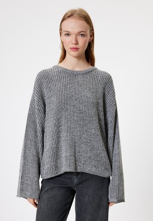 Grauer Strickpullover mit einer dicken Ripptextur, weiten Ärmeln und einem runden Halsauschnitt. Kombiniert mit dunkelgrauen, hoch taillierten Jeans.
