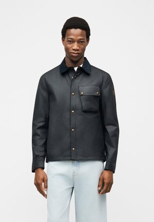 Ung mand iført en mørk knappet jakke med brystlomme og lyseblå jeans, stående mod en ensartet lys baggrund.