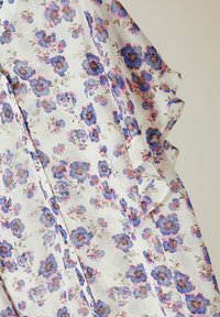 Blumenbluse aus leichtem weißen Stoff, mit blauen und rosa Blumen, Rüschenschößen und einer Knopfdetaillierung vorne.