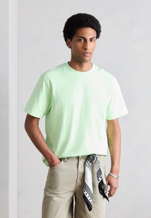 Νέος άνδρας με κοντά σγουρά μαλλιά φοράει ανοιχτοπράσινη μπλούζα και μπεζ παντελόνι με ένα ασπρόμαυρο μαντήλι προσαρτημένο, στέκεται με το ένα χέρι στην τσέπη.