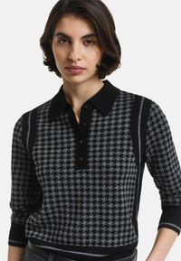 Vrouw met kort donker haar, gekleed in een zwart-grijze houndstooth gepatterneerde langarmige poloshirt met een zwarte kraag en knopen.