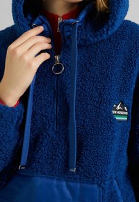 Modrá fleecová mikina s texturovaným povrchem, nastavitelnými šňůrkami, zapínáním na zip a kontrastním modrým spodním lemem s detailem loga.