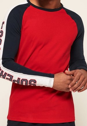 Mand iført en rød langærmet skjorte med marineblå ærmer med hvide striber og teksten "SUPER" på venstre arm, hænderne foldet foran.