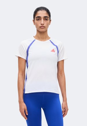 Femme aux cheveux plaqués en arrière portant un t-shirt de sport blanc avec des rayures bleues et des leggings bleus, debout devant un fond uni.