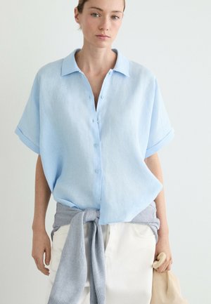 Femme portant une chemise bleu clair à manches courtes boutonnée, un pull gris noué à la taille, un pantalon blanc, tenant la poignée d'un sac beige sur un fond uni.
