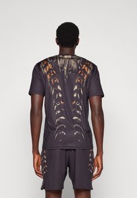 Man som står med ryggen mot kameran, iförd matchande svarta skjorta och shorts med abstrakt mönster av palmer i orange och beige på baksidan och sidorna.