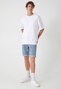 Biała bawełniana koszulka z krótkim rękawem, połączona z jasnoniebieskimi dżinsowymi szortami i białymi sneakersami. Stylizacja charakteryzuje się luźnym krojem i prostym designem.