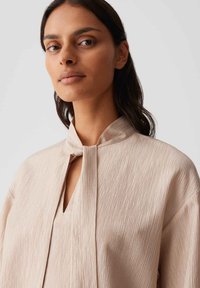 Beige blouse with a textured finish, open round neckline, and a high collar design. Features a keyhole opening at the neck.