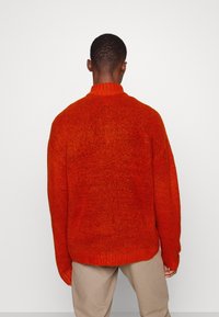 Roter gestrickter Pullover mit hohem Kragen, strukturiertem Stoff und oversized Passform, von hinten gezeigt, kombiniert mit hellbeigen Hosen.