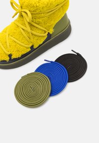 Žlutá boot s fleece podšívkou a zelenými koženými akcenty, s kulatými šňůrkami v olivové, modré a černé barvě, vystavená na bílém pozadí.