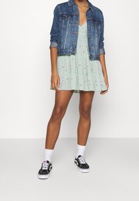 Ljusgrön blommig klänning med volangkant, i kombination med en blå denimjacka. Svart och vit sneakers med vita ankelsockor.