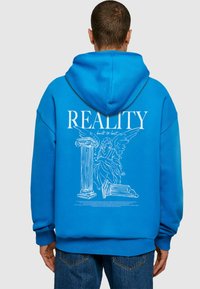 Modrá mikina s kapucňou s veľkým bielym grafick�ým potlačou, na ktorej sú anjel a stĺpy, text "REALITY built to last" nad nimi a zložitými čiarovými detailmi.