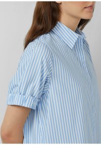 Camicia a righe azzurro chiaro e bianca con colletto pieghevole e maniche corte arrotolate. Realizzata in un tessuto liscio con un design classico.