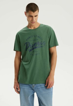 Młody mężczyzna w zielonej koszulce z logo "Diverse" i jasnoniebieskich dżinsach, patrzy w dół z neutralnym wyrazem twarzy.