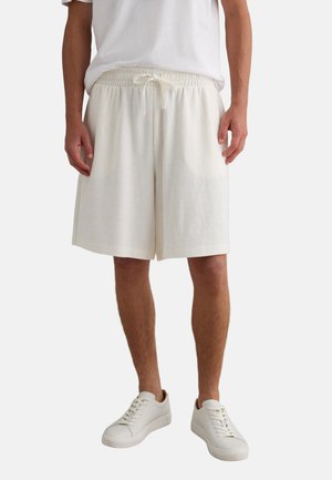 Hombre vistiendo pantalones cortos de cordón color blanco hueso, camiseta blanca y zapatillas blancas, de pie contra un fondo claro liso.