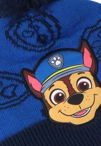Beanie lavorato a maglia blu con il volto di un cane cartoon con un cappellino, motivo testurizzato e un pom-pom soffice in cima.