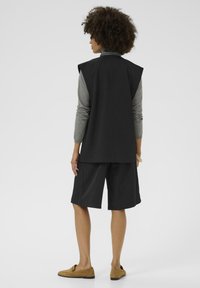 Black sleeveless vest over a gray long-sleeve top, paired with black shorts, and light brown slip-on shoes. Plain background.