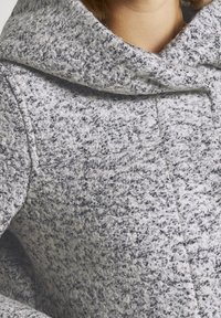 Texturovaný šedobílý vlněný kabát s velkou kapucí, se zapínáním na knoflík a viditelnými stehy. Měkký povrch s útulným vzhledem.