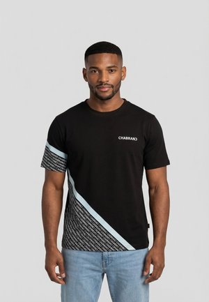 Hombre vistiendo una camiseta negra de Chabrand con patrones diagonales en azul claro y logos repetidos, combinada con jeans azul claro, de pie mirando hacia adelante.