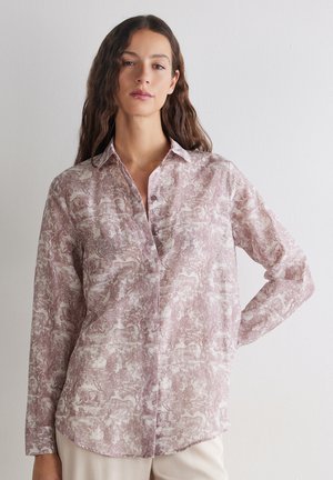 Kvinde med langt bølget hår iført en lys mauve skjorte med knapper og et dyre- og naturmønster, poserer med den ene hånd på hoften mod en ensfarvet baggrund.