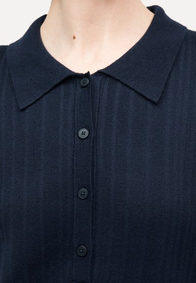 Κοντινή λήψη ενός ατόμου που φοράει μπλούζα σκούρου ναυτικού με ριμπ ύφανση, γιακά πόλο και τέσσερα μαύρα κουμπιά στο μπροστινό μέρος.