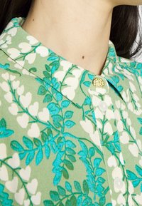 Grün karrierte Hemd mit einem floralen Muster in Weiß und Türkis, strukturiertes Material und einem dekorativen goldenen Knopf.