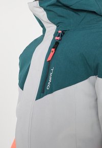 Chaqueta en tonos verde azulado y gris con un cuello alto, cierre frontal de cremallera y detalles en naranja en contraste, con el nombre de la marca "O'Neill" destacado.