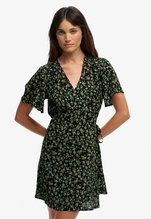 Femme aux longs cheveux bruns portant une robe portefeuille noire à fleurs avec des manches courtes volantées, debout les mains sur les hanches.