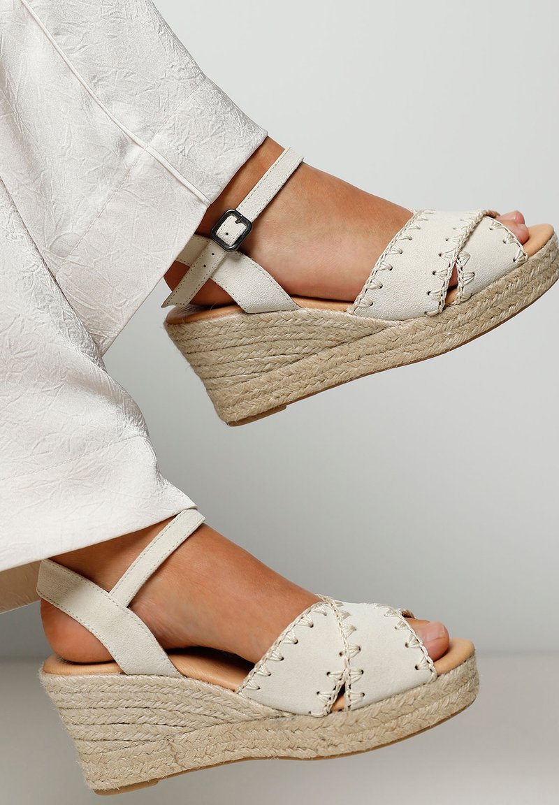 Wedge sandals with a light beige suede upper, crisscross straps, ankle strap with a metal buckle, and a textured jute-wrapped platform.