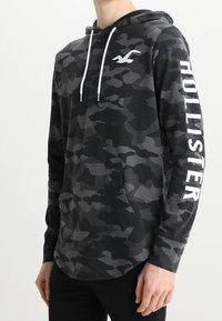 Svart och grå camouflagetröja med framficka, vita dragsnören och "HOLLISTER" tryckt med stora vita bokstäver på ärmen.
