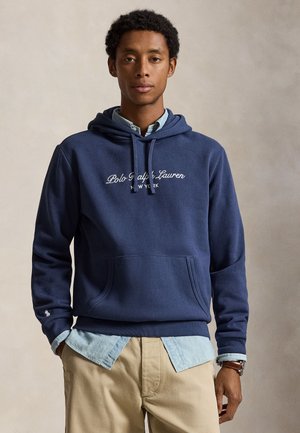 Un homme porte un sweat à capuche bleu marine Polo Ralph Lauren par-dessus une chemise bleu clair et un pantalon kaki, sur un fond beige uni.
