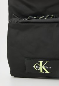 Svart nylon ryggsekk med en glatt tekstur, glidelåslomme med en svart og grønn snor, og et logomerke med "Calvin Klein" i hvitt og grønt.