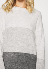 Femme portant un pull en tricot à manches longues avec des blocs de couleur horizontaux en blanc, gris clair et gris foncé, cheveux blonds et ondulés.