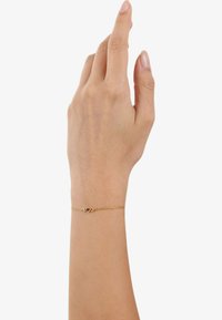 Bracciale d'oro con due pietre decorative rosse, collegate da una delicata catena, indossato su una mano con smalto neutro.