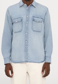 Camisa de mezclilla azul claro con mangas largas, cierre de botones, dos bolsillos en el pecho y cuello clásico puntiagudo. Tela texturizada con un ligero brillo.