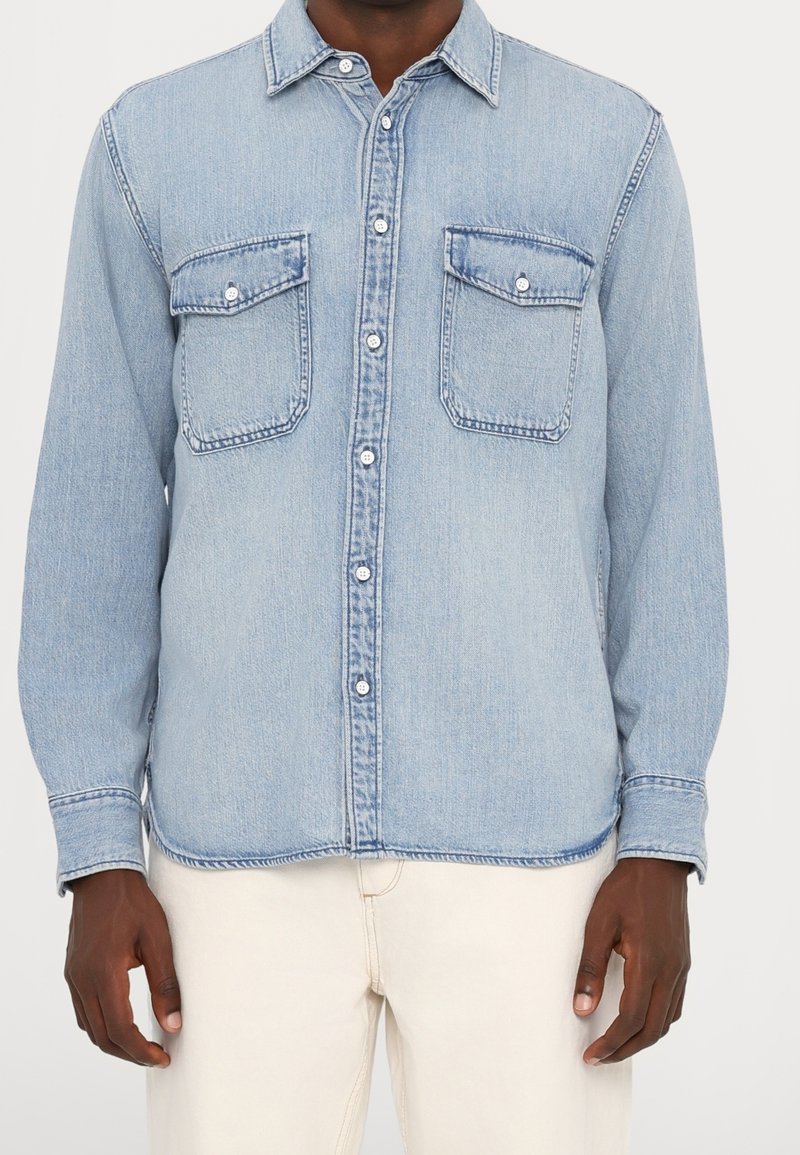 Camisa de mezclilla azul claro con mangas largas, cierre de botones, dos bolsillos en el pecho y cuello clásico puntiagudo. Tela texturizada con un ligero brillo.