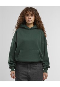 Grønn hettegenser med stor frontlomme, oversized design og snorhette. Har ribbestrikkede mansjetter og en myk tekstur. Båret med mørke grå jeans.