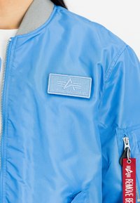 Modra bombarderska jakna iz lahkega materiala, zivo sivo rebrastim ovratnikom, oznako blagovne znamke na prsih in rdečim trakom na zadrgi.