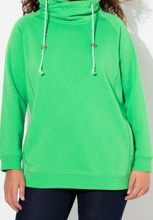 Persona que lleva una sudadera con capucha verde brillante con cordones blancos y verdes y pantalones azul oscuro, de pie frente a un fondo blanco.