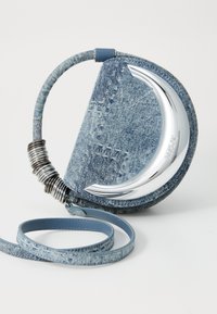 Geantă crossbody rotundă din denim, cu detaliu metalic în formă de semilună argintie și inele metalice înfășurate pe mâner, pe fundal alb.