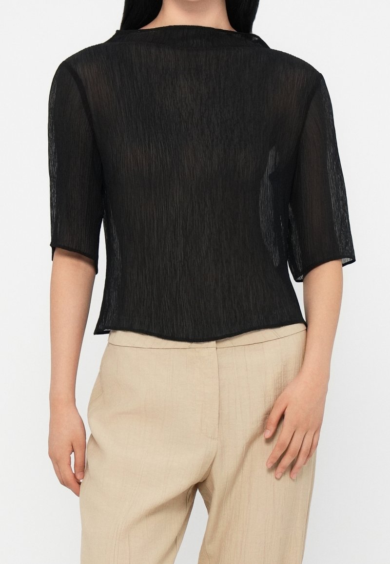 Haut noir transparent à manches courtes, présentant un design texturé froissé et un col rond. Porté avec un pantalon beige clair.