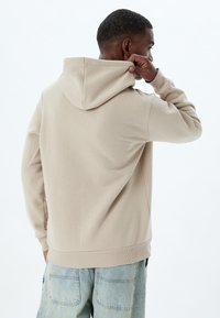 Sudadera con capucha beige de textura suave, con puños y cintura acanalados. Presenta un corte relajado y capucha con cordón.