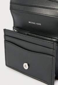 Juodas odinis kortelių dėklas, turintis kelias kortelių kišenes, užtrauktuku užsegamą skyrių ir sidabrinį spaudžiamą mygtuką. Matoma Michael Kors prekės ženklo etiketė.