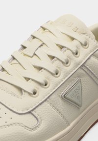 Αθλητικό παπούτσι Guess σε κρεμ απόχρωση με δερμάτινη υφή, κρεμ κορδόνια και ασημένιο τριγωνικό λογότυπο Guess στην πλευρική επιφάνεια.