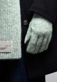 Svart kappa med en ljusgrön ribbad strumpa. Handgelen har en etikett med texten "PARADISE STREET ONE." Knappdetalj syns på kappan.