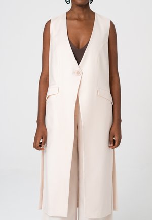 Długa, bezrękawnikowa kamizelka w kremowym kolorze z zapięciem na jeden guzik, noszona na ciemnobrązowym topie z dekoltem w serek przez stojącą osobę.