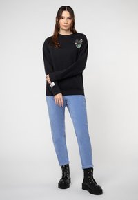 Schwarzer Hoodie mit Grafikdruck auf der linken Schulter, kombiniert mit hellblauen Cordhosen und schwarzen Schnürstiefeletten.