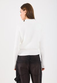 Fehér kötött pulóver bolyhos textúrával és bordázott ujjakban, sötét, enyhén kopott farmernadrággal párosítva, melyek hátul zsebekkel rendelkeznek.