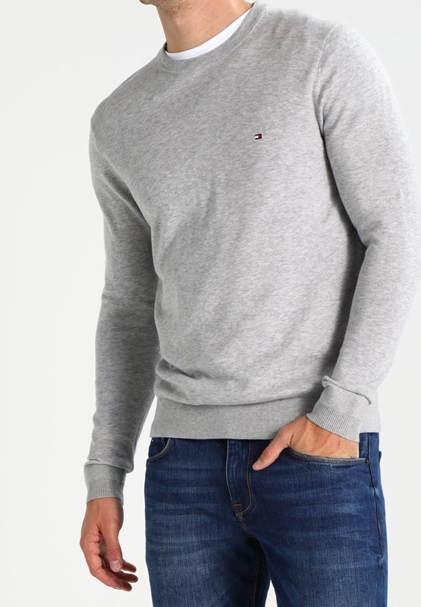 Homme portant un pull gris clair à col rond et un jean bleu, une main dans la poche, sur fond blanc uni.